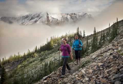 Two women smiling on their hike along a scuree path with low hanging clouds around mountains in the background.