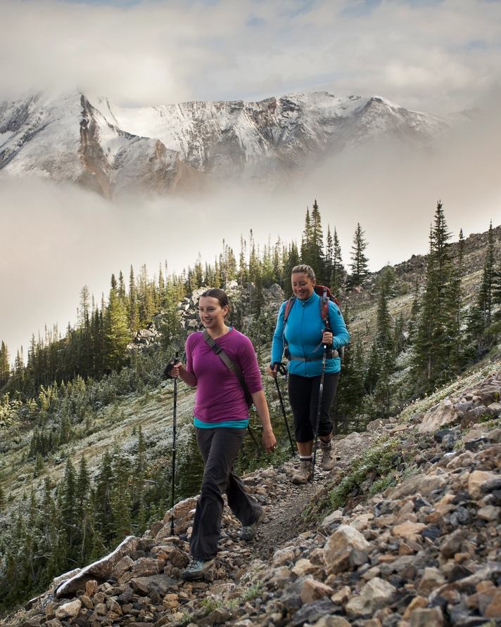 Two women smiling on their hike along a scuree path with low hanging clouds around mountains in the background.