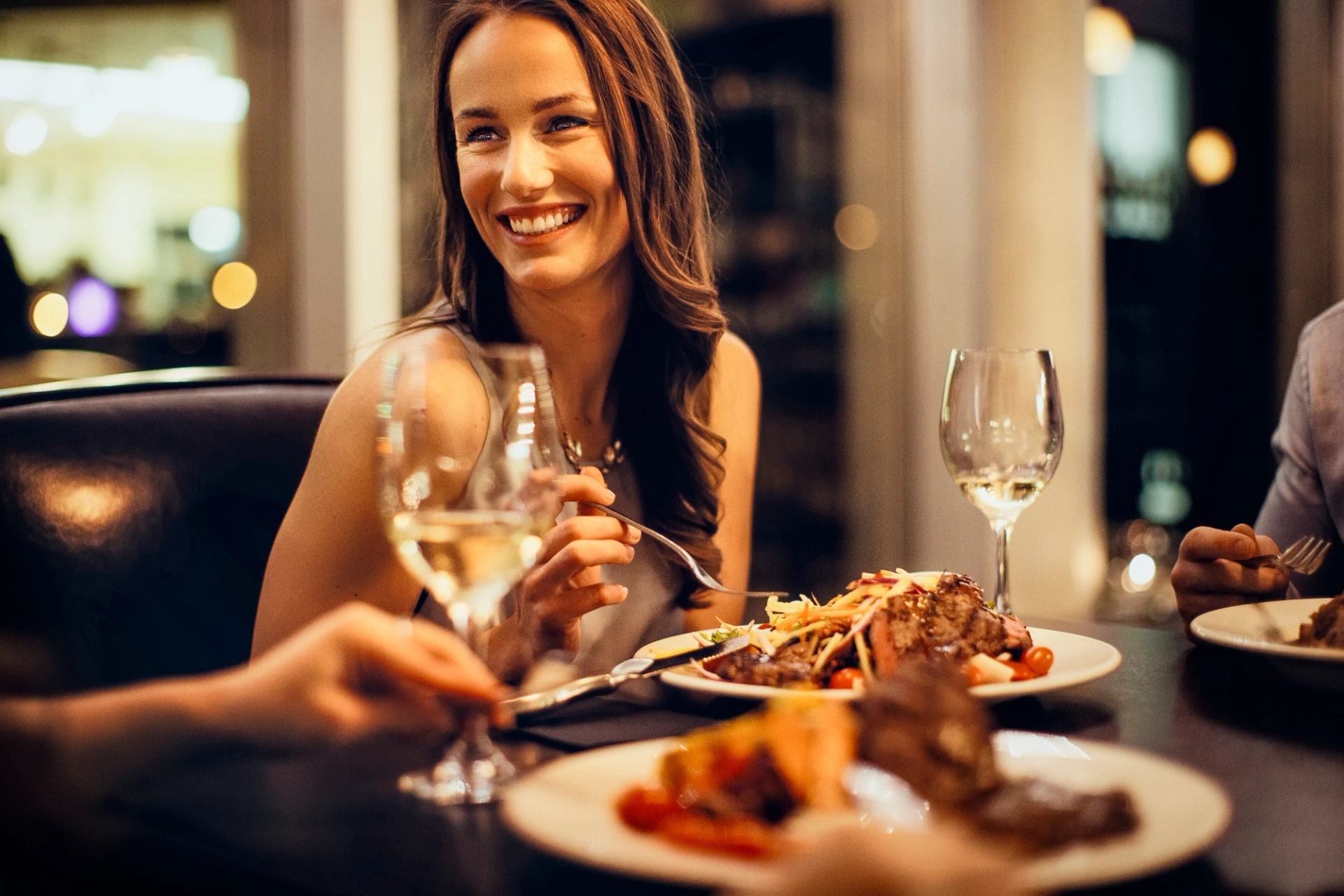 Closeup of a woman dining with a plate of food and a glass of wine on the table in front of her.