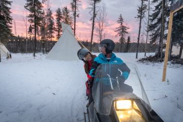 A couple smiling at each other on a snowmobile at sunset with a tipi in the background.