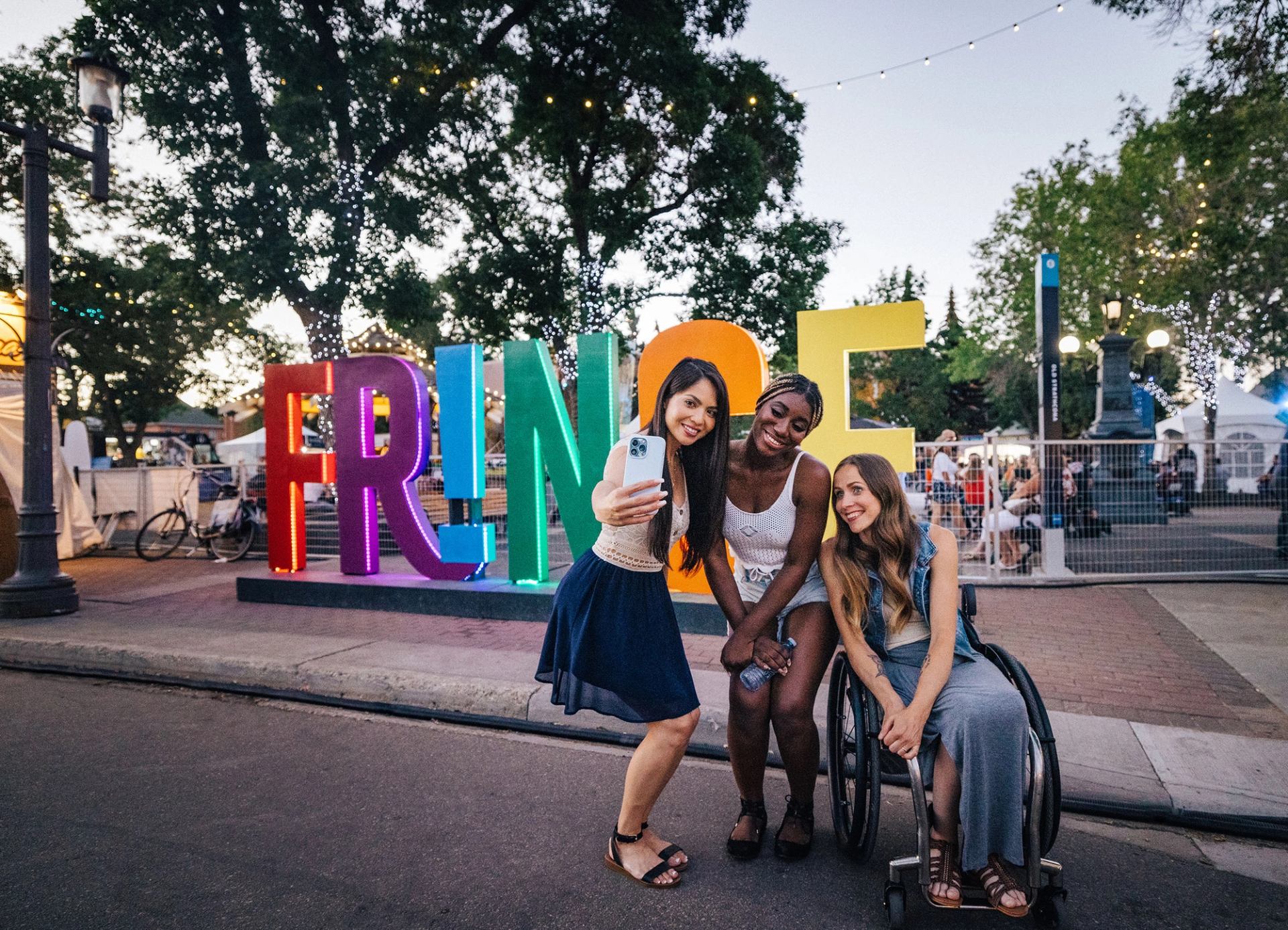 Three women, one in a wheelchair, take a selfie in front of a large sign that says Fringe.