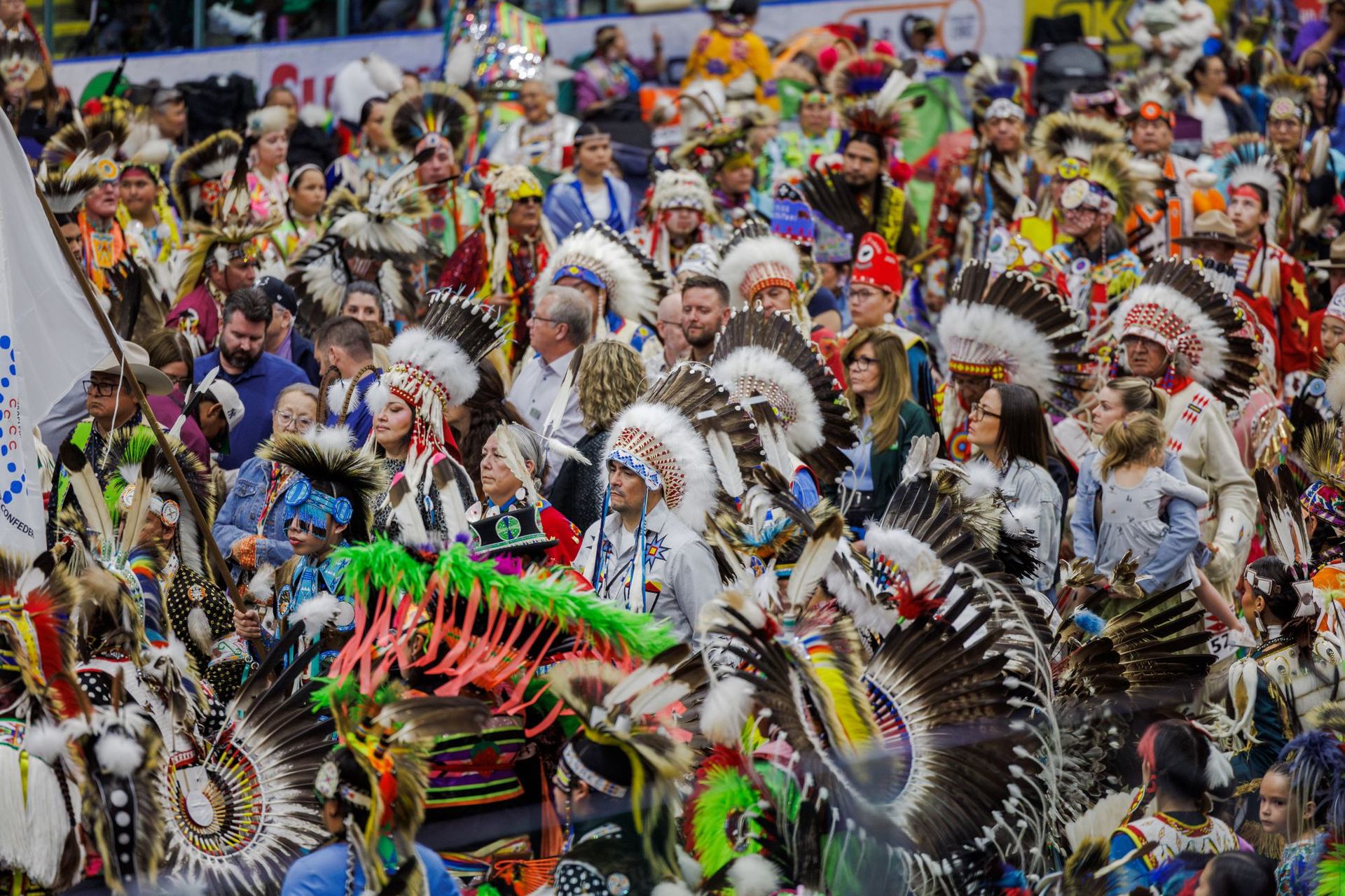 A large, colourful crowd at a powwow, featuring dancers, singers, chiefs and attendees.