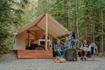 Campers sit by a campfire roasting marshmallows in front of a cabin in the trees.