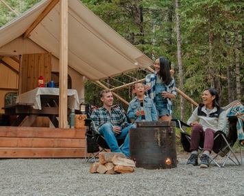 Campers sit by a campfire roasting marshmallows in front of a cabin in the trees.