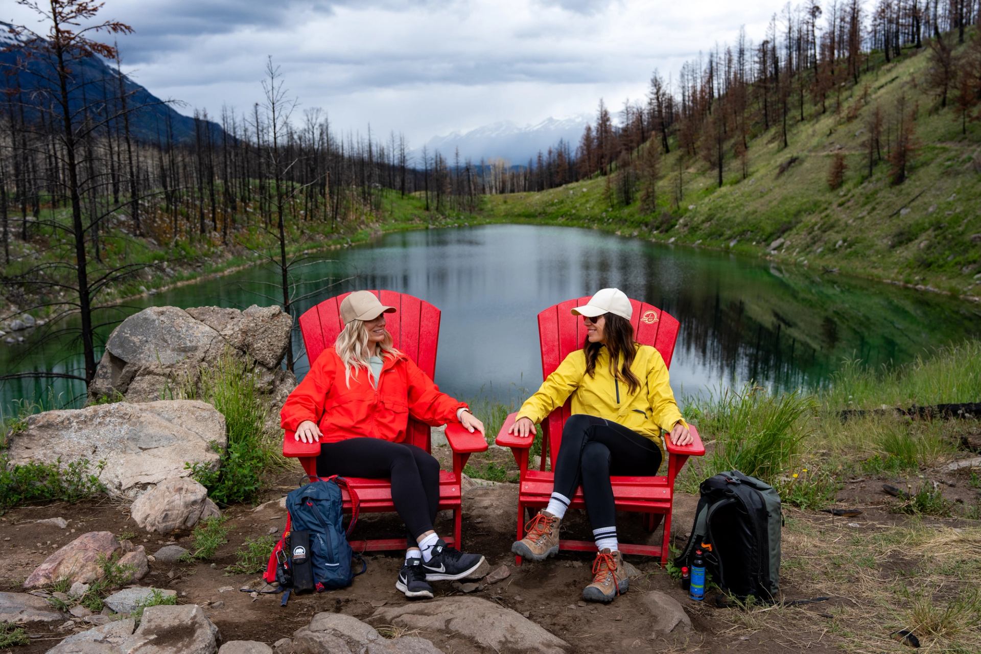 Two hikers sit in red Adirondack chairs on a lakeshore in Jasper National Park with a regenerating forest surrounding them.