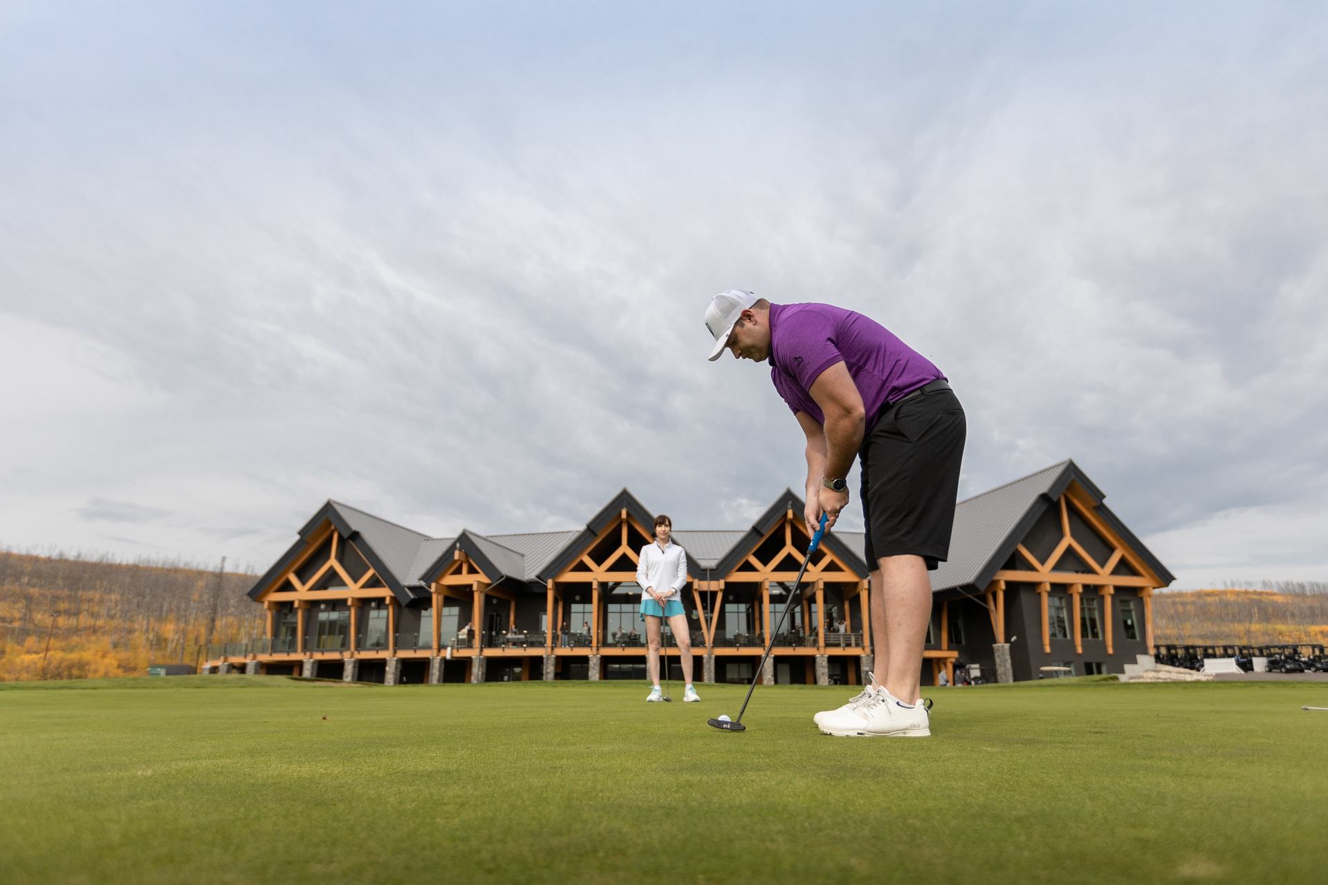 Man putting on green woman behind watching with the Fort McMurray Golf Club in the background.