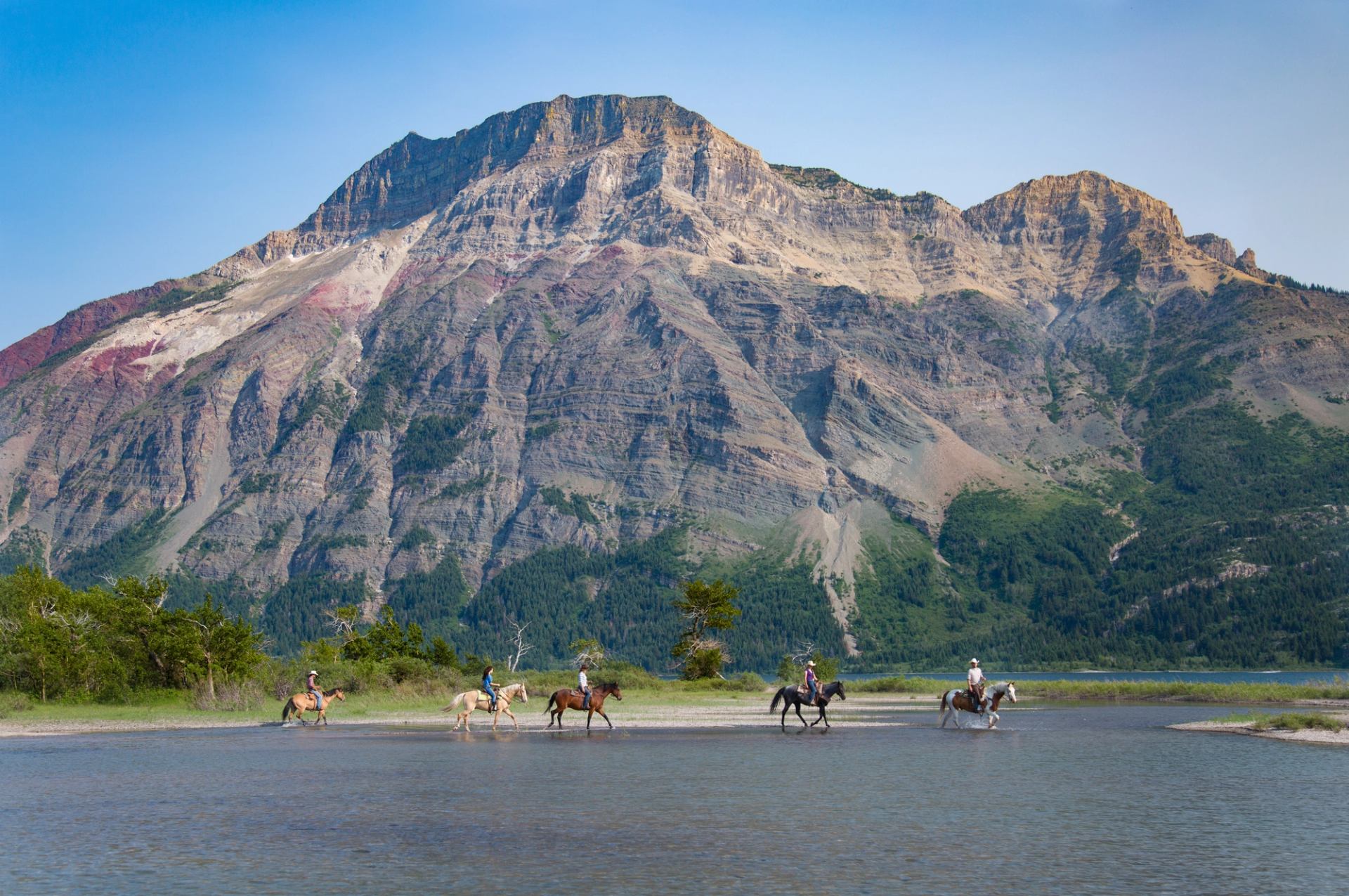 People riding horses on a lake shoreline with the Rocky Mountains in the background.