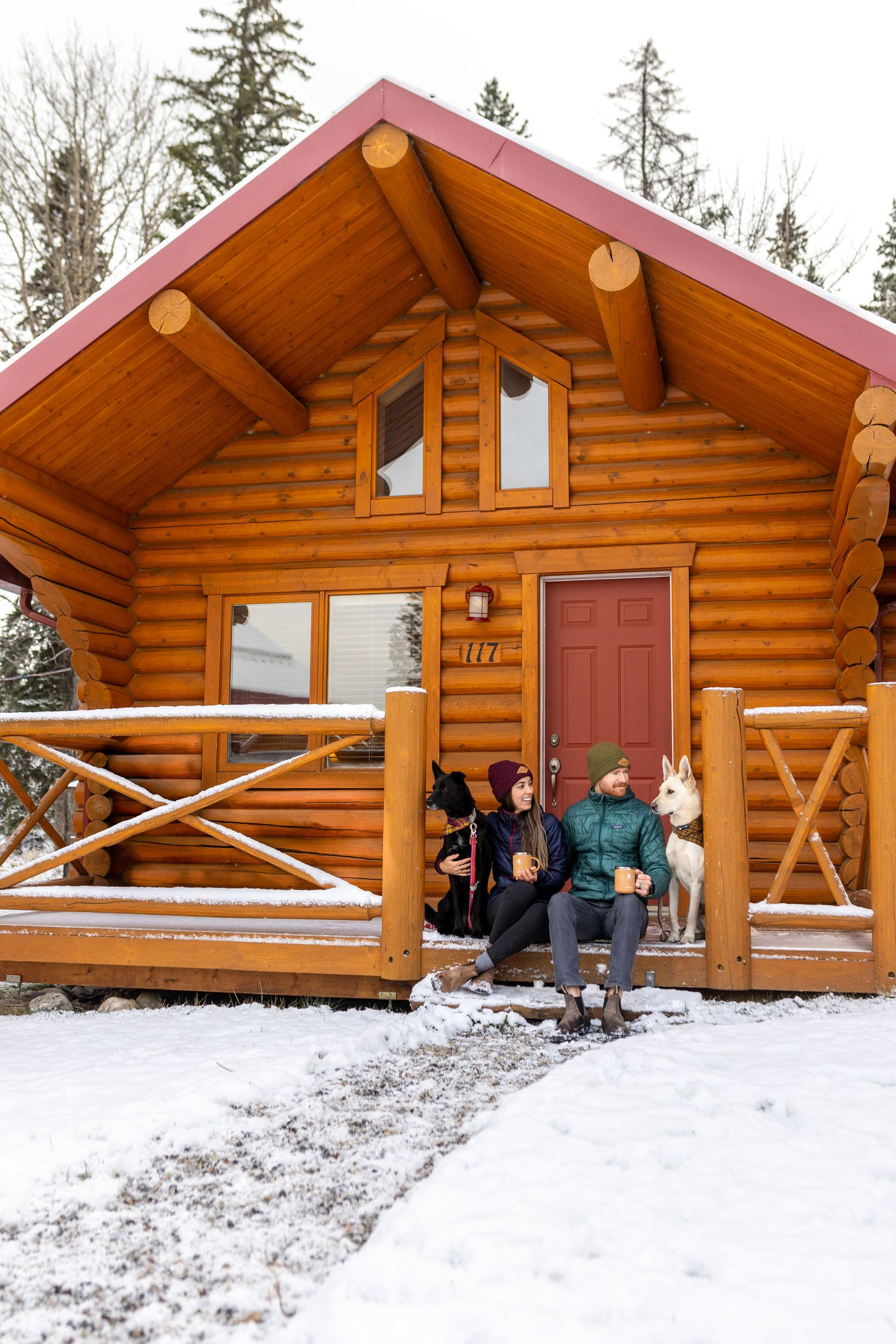 A young couple with two dogs sit on porch of Miette Mountain Cabins during winter while drinking hot chocolates.