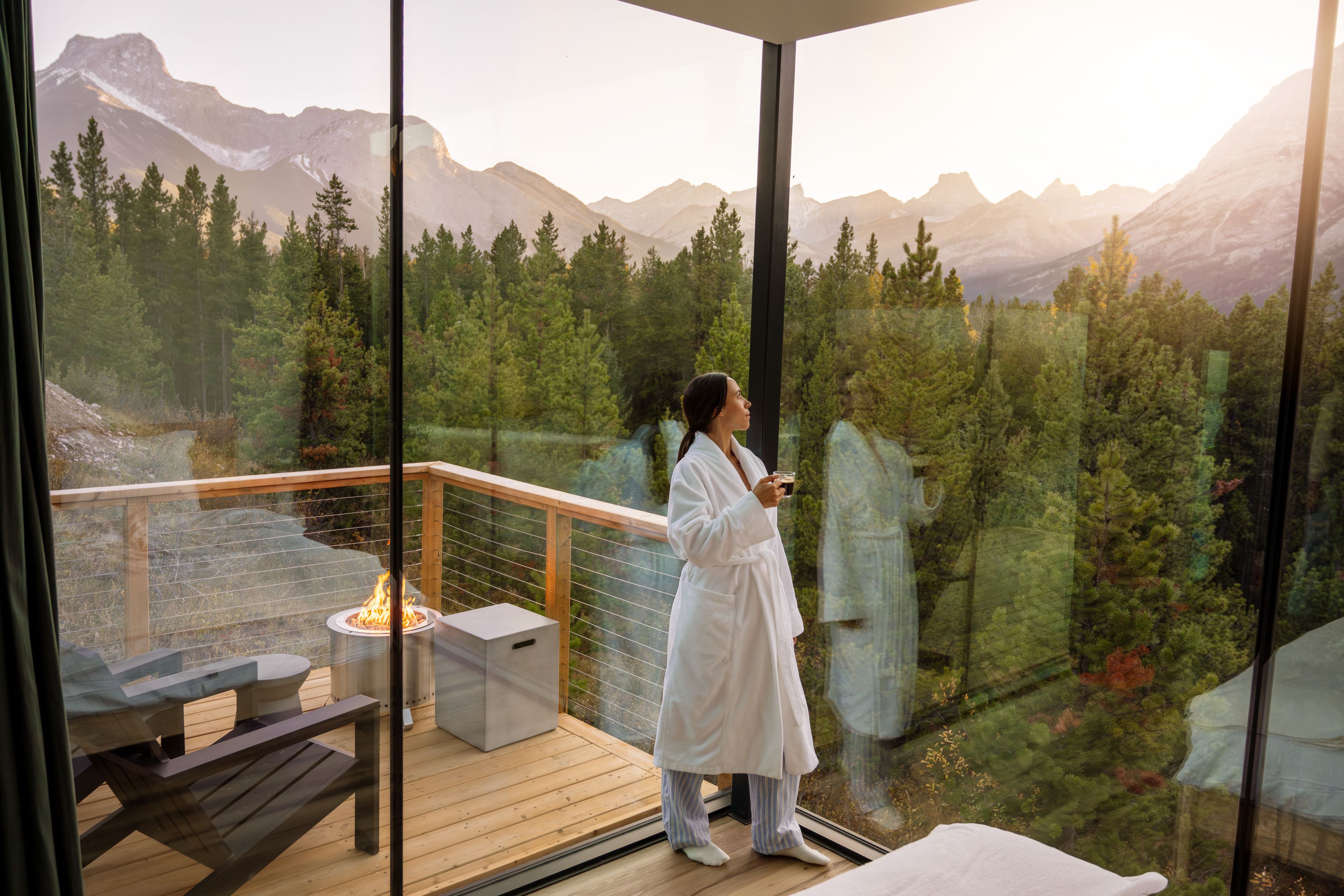 A woman stands against the glass walls of her cabin, looking out at the Canadian Rockies at Skyridge Glamping in Alberta.