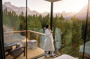 A woman stands against the glass walls of her cabin, looking out at the Canadian Rockies at Skyridge Glamping in Alberta.