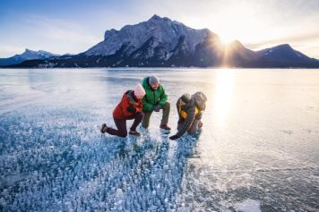 A guide and 2 guests look at the Abraham Lake ice bubbles with the Rockies in the distance.