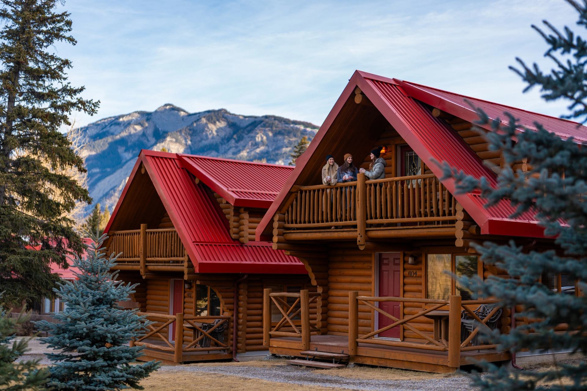 Three friends stand on the second-floor balcony of their red-roofed cabin in Jasper National Park