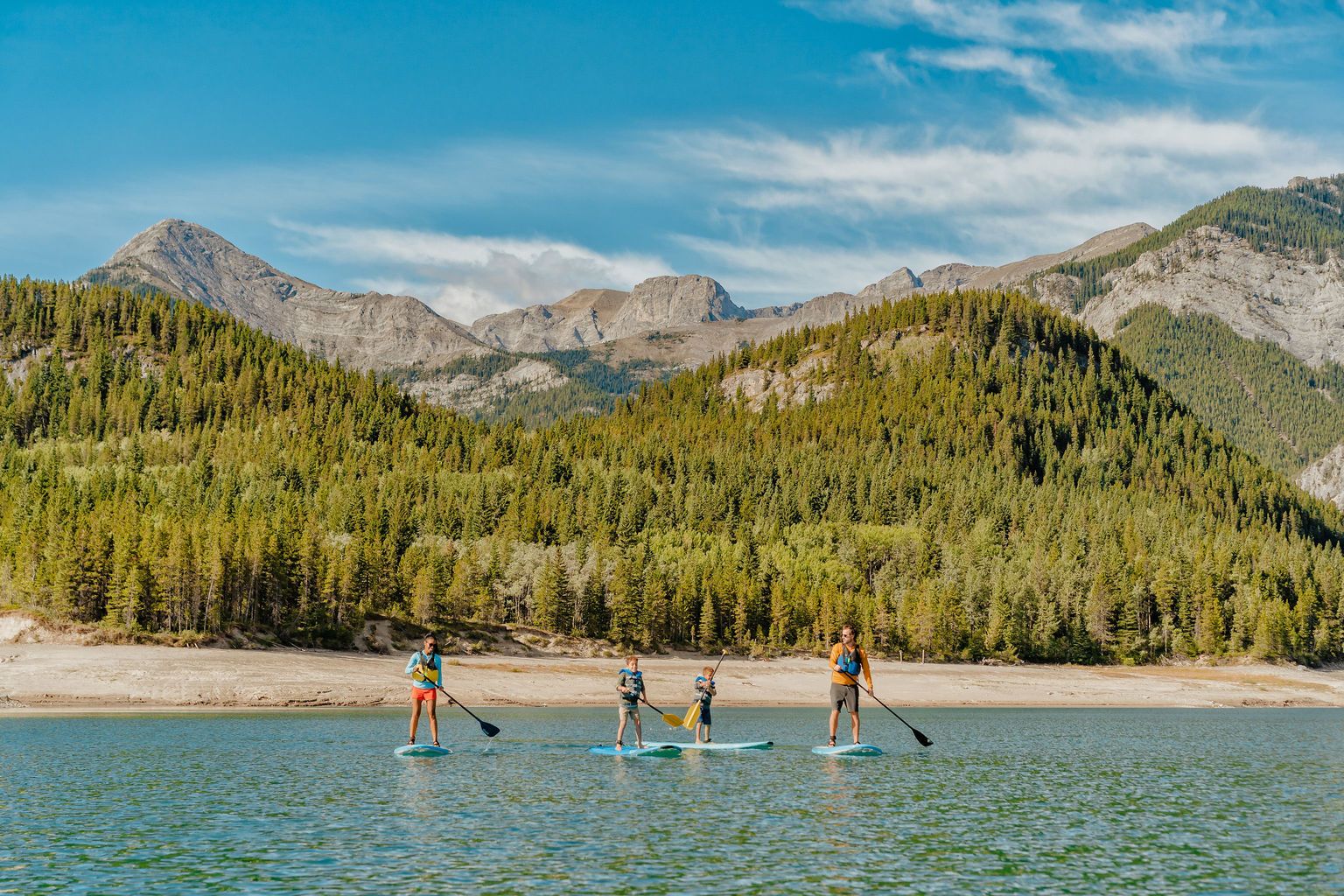 People paddleboard on a lake with a beach, trees and mountains in the background.