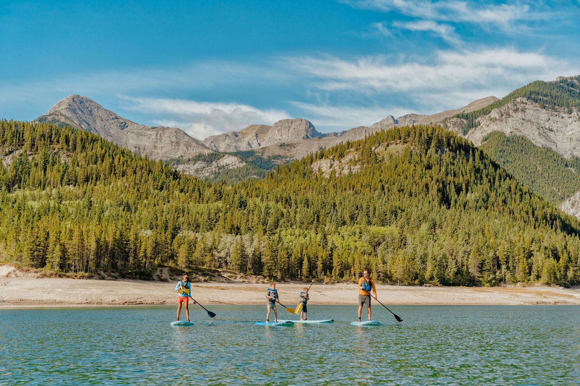 People paddleboard on a lake with a beach, trees and mountains in the background.