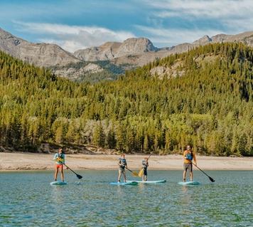 People paddleboard on a lake with a beach, trees and mountains in the background.