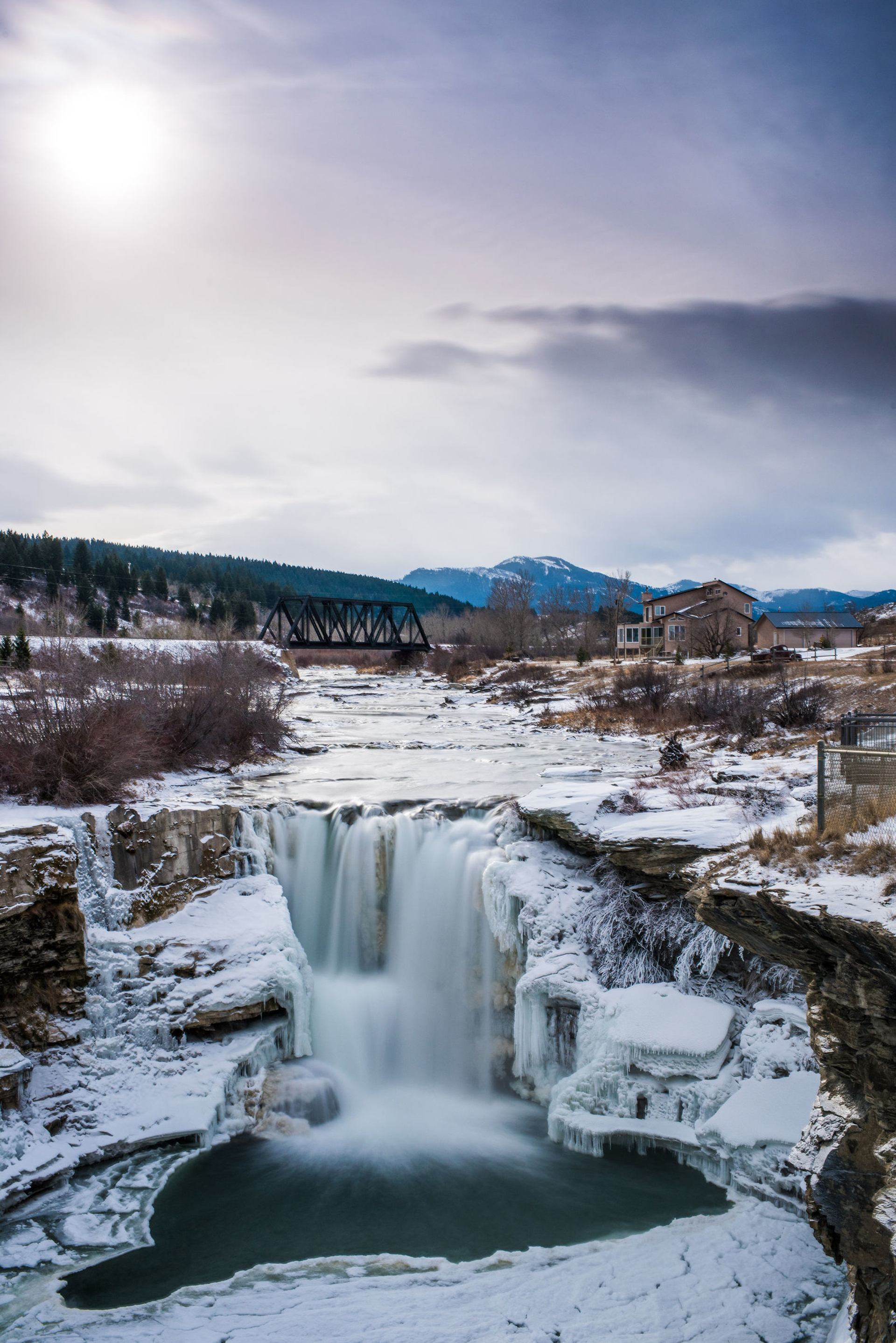 Aerial view of waterfalls in winter with a bridge and mountains in the background.