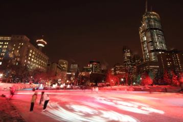 People ice skating at night on an outdoor rink in a lit downtown plaza.