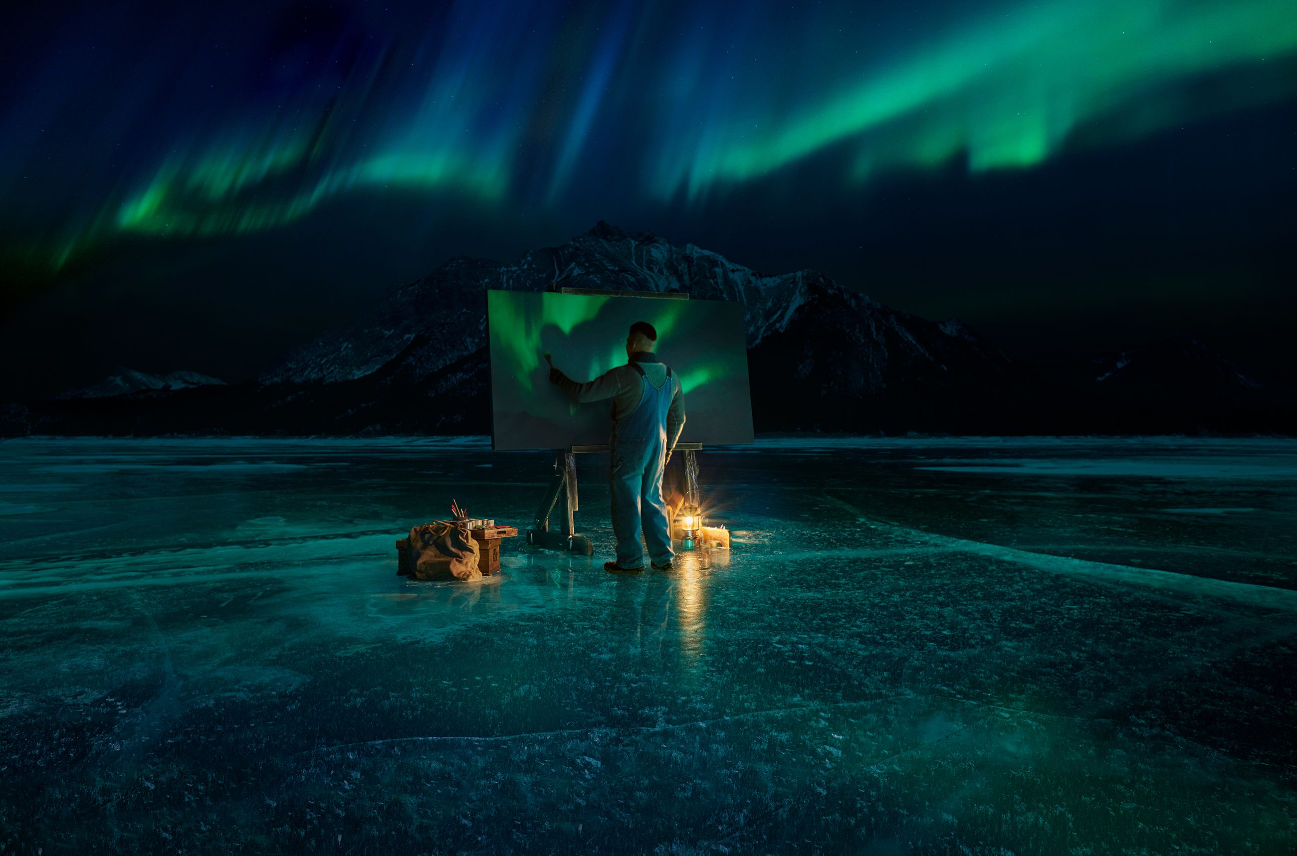 Man standing on frozen lake at night painting the Northern Lights on a large canvas with the Northern Lights overhead in the sky.