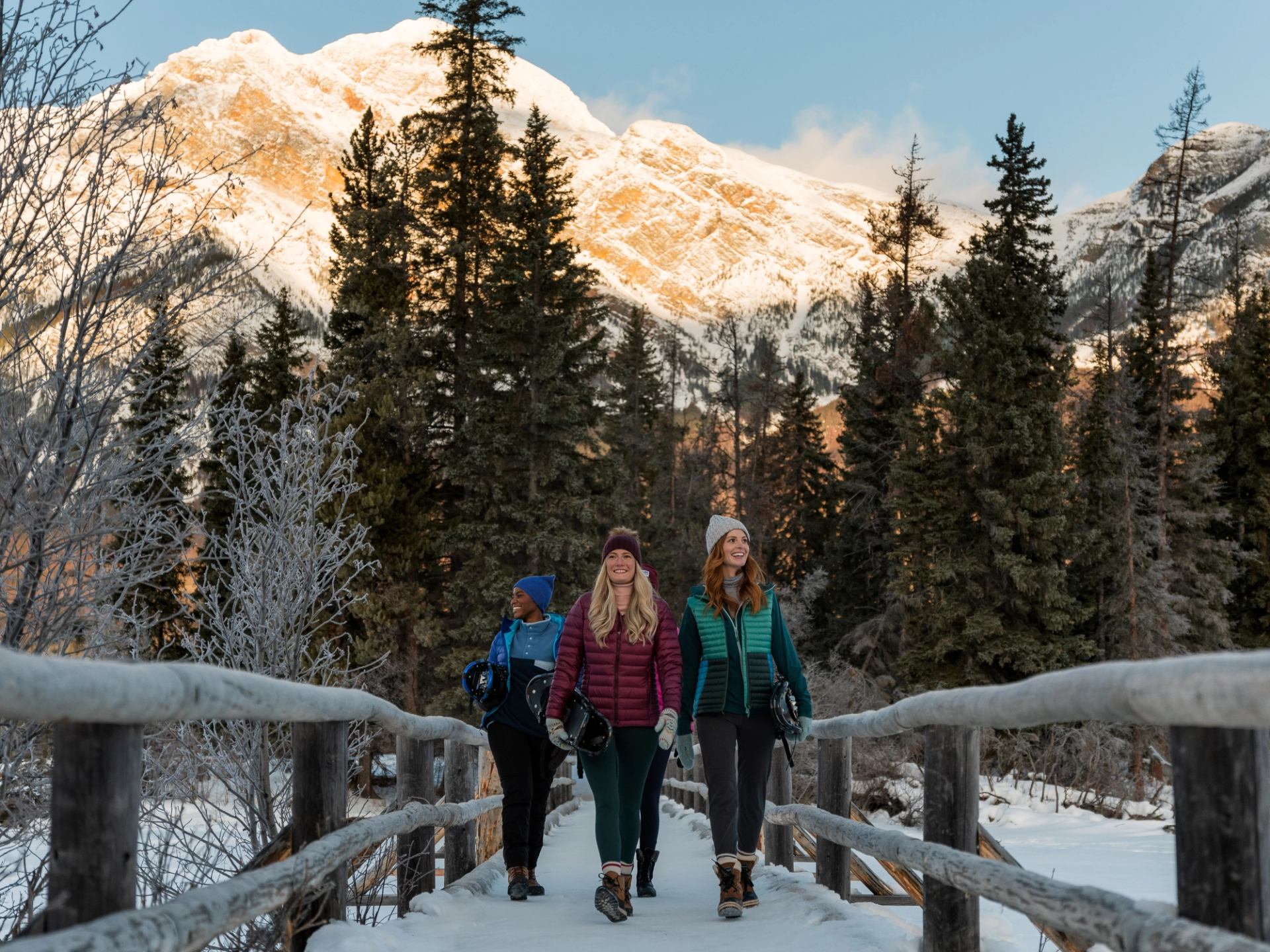 Four friends cross a bridge in the Canadian Rockies carrying snowshoes on a pleasant winter day.