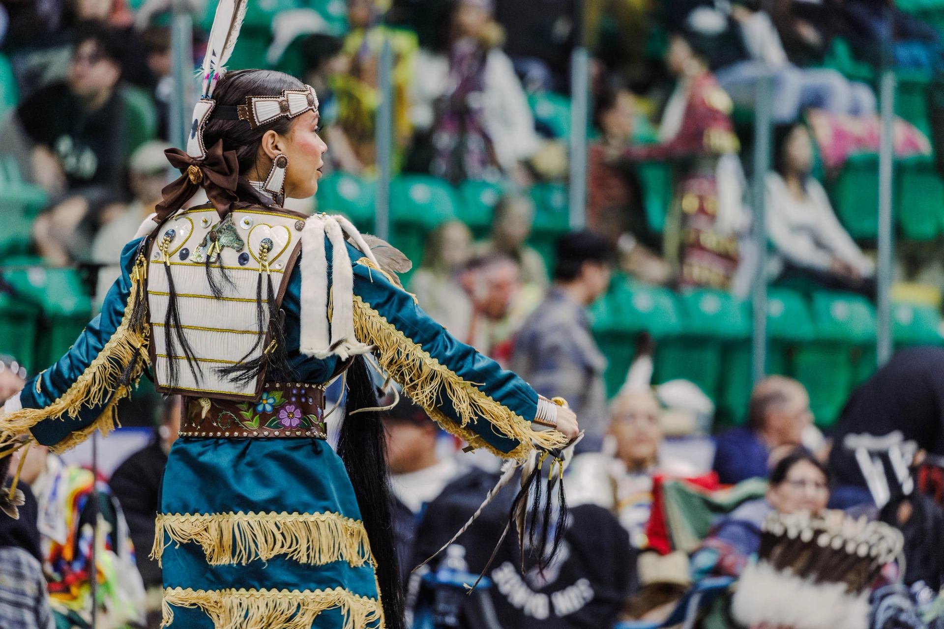 A female dancer in deep turquoise regalia trimmed with gold performs during a powwow.