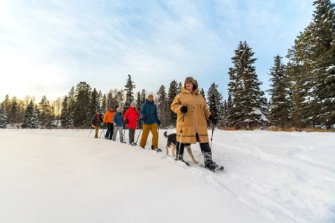 Indigenous Knwoledge Keeper Brenda Holder continues to guide the group on the snowshoe walk across the lake with blue skies and trees in the background.