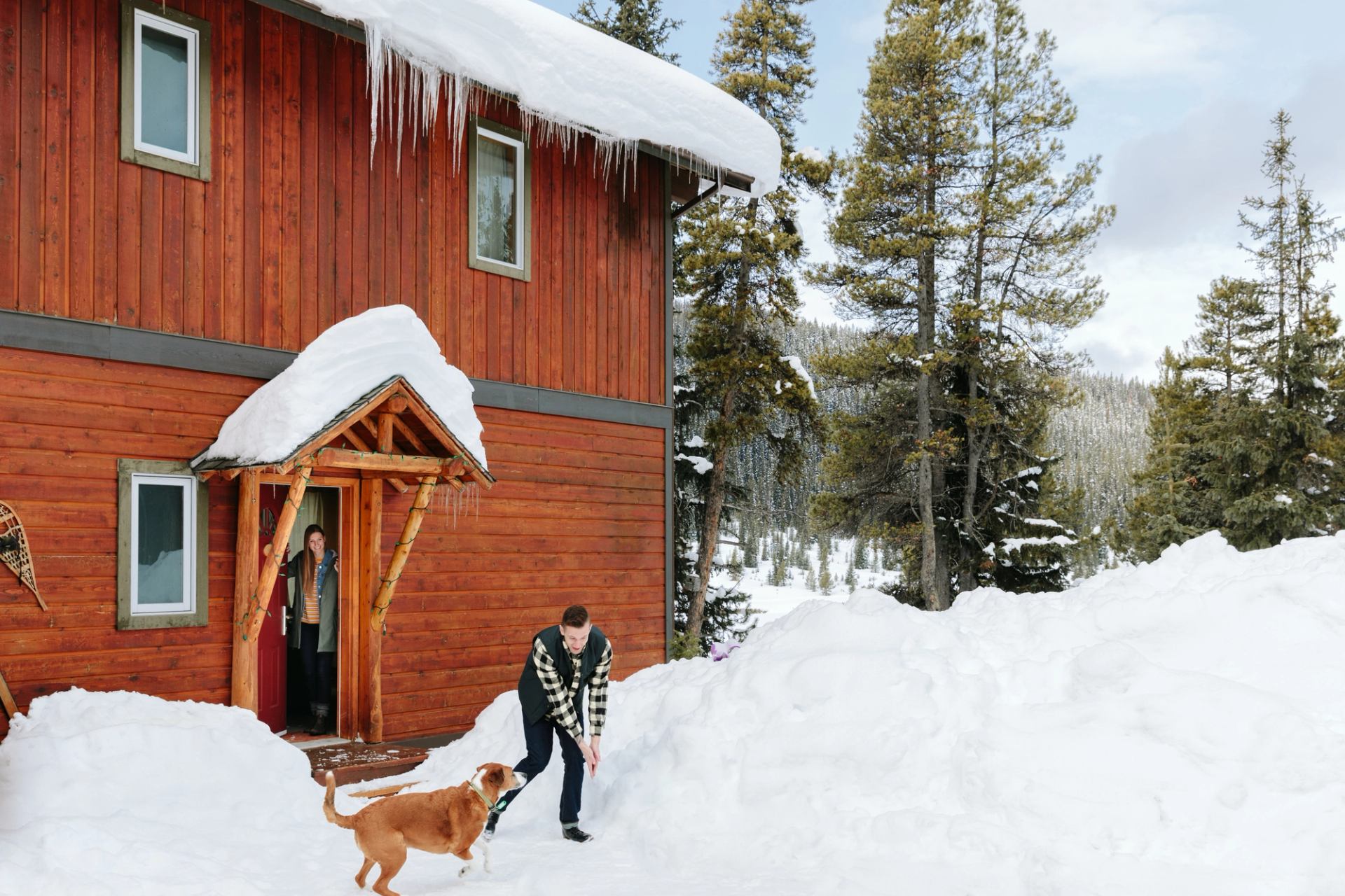 Couple playing with a dog in the snow outside a log lodge.