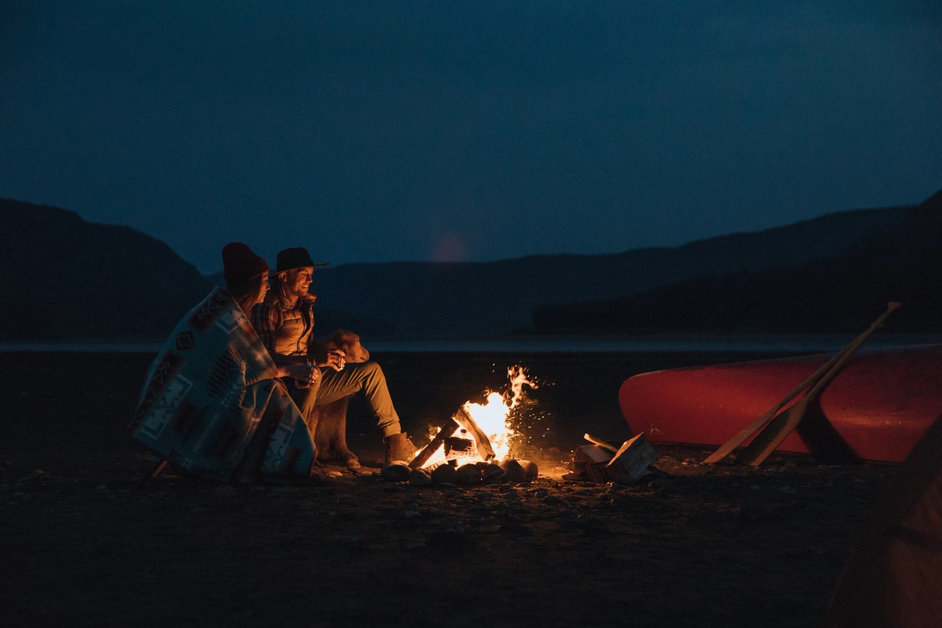 Two people and a dog camping and sitting by the campfire with a canoe and lake in the background.