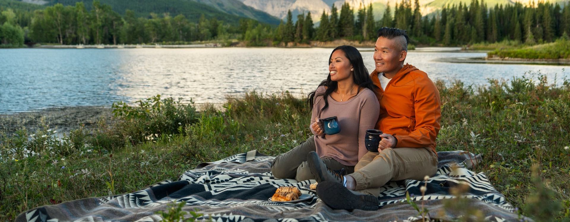 A couple sits by the water taking in sunrise at Glamping Resorts in Castle Provincial Park while sipping on coffee and enjoying pastries for breakfast.