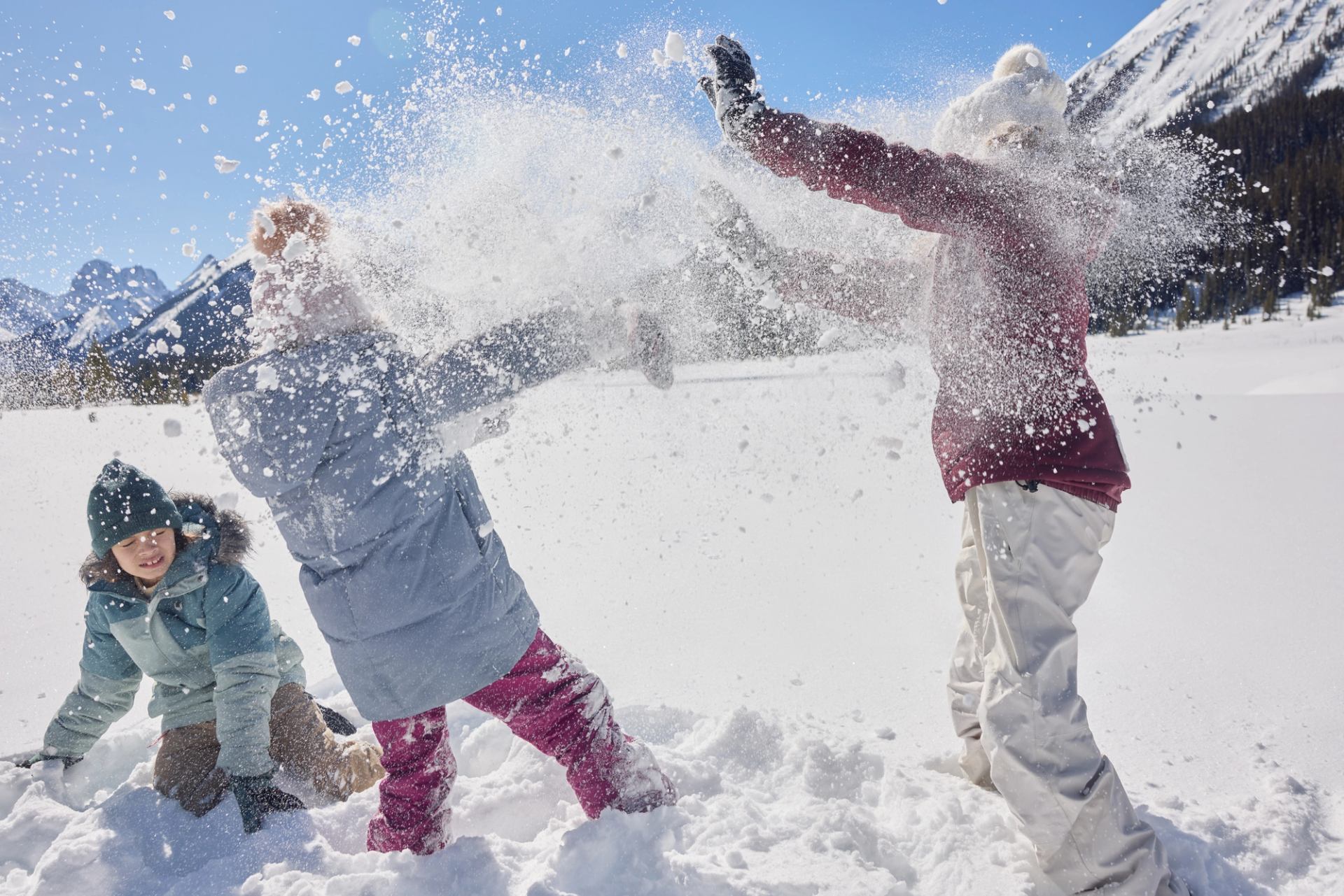Three kids have a snowball fight in a mountain setting on a blue-sky day.