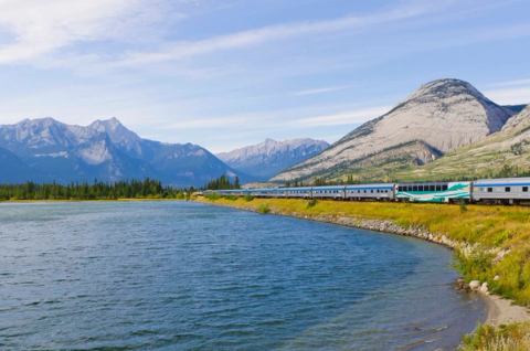 VIA Rail train travelling along a pristine lake on a sunny day as it enters the Rock Mountains towards Jasper.