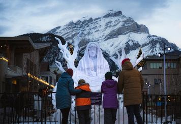 A family looking at a snow sculpture with mountains in the background.