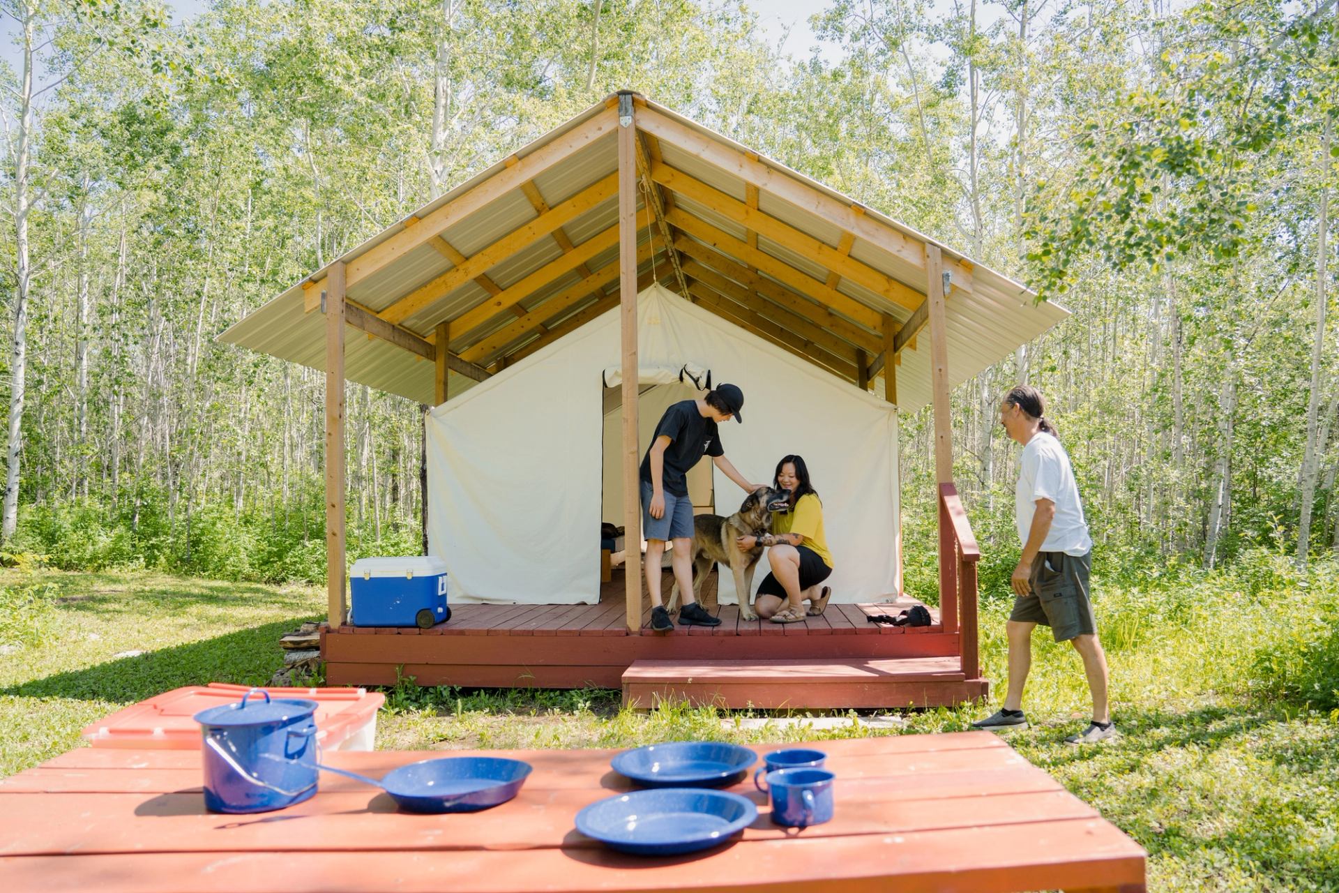 A woman and a teenage boy pet a dog on the deck of a small wooden cabin in the woods, with a man standing nearby.