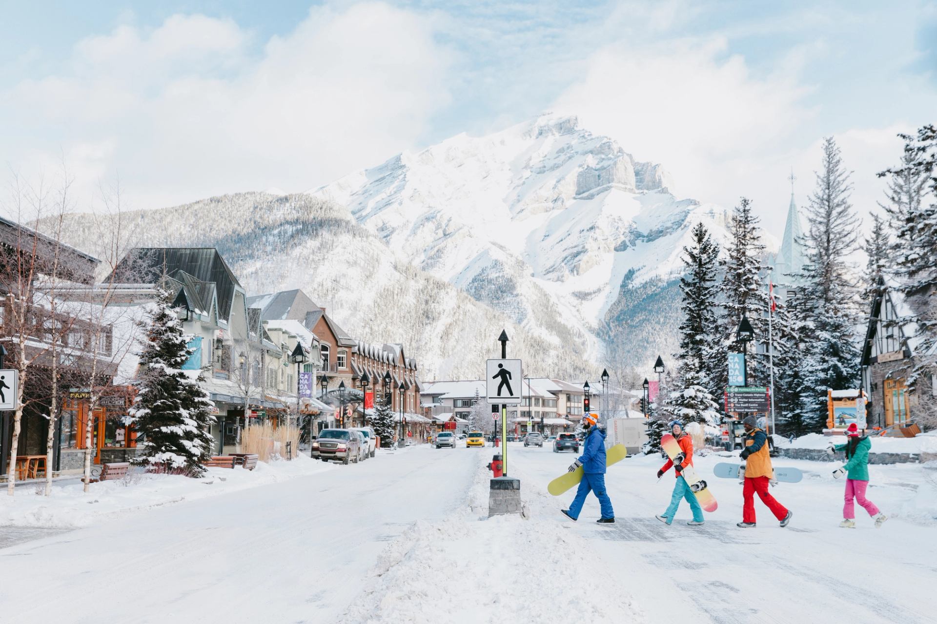 A group of skiers and snowboarders walking across Banff Avenue, significant mountain view in the background, in Banff National Park