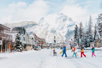 A group of skiers and snowboarders walking across Banff Avenue, significant mountain view in the background, in Banff National Park