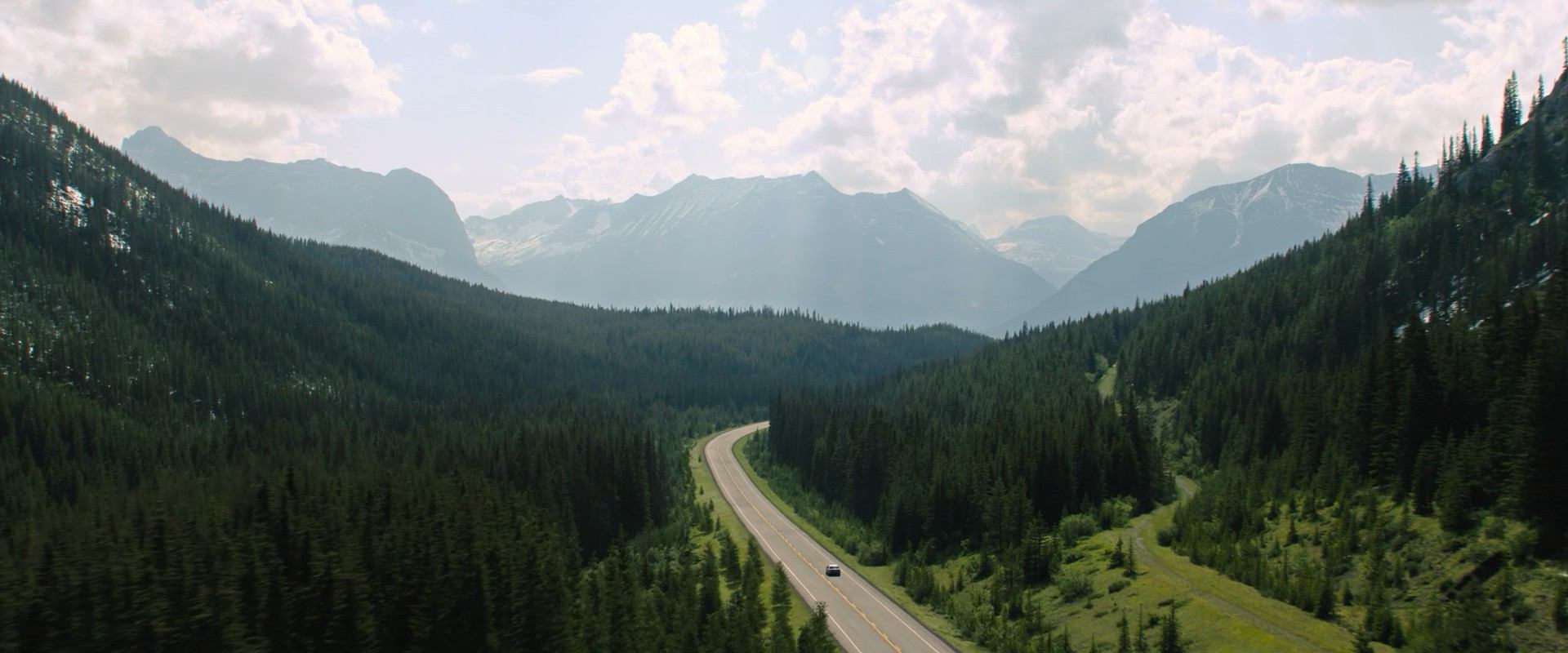 A movie still from Reminders of Him featuring green mountain foothills and distant rocky mountains with a highway through it all.