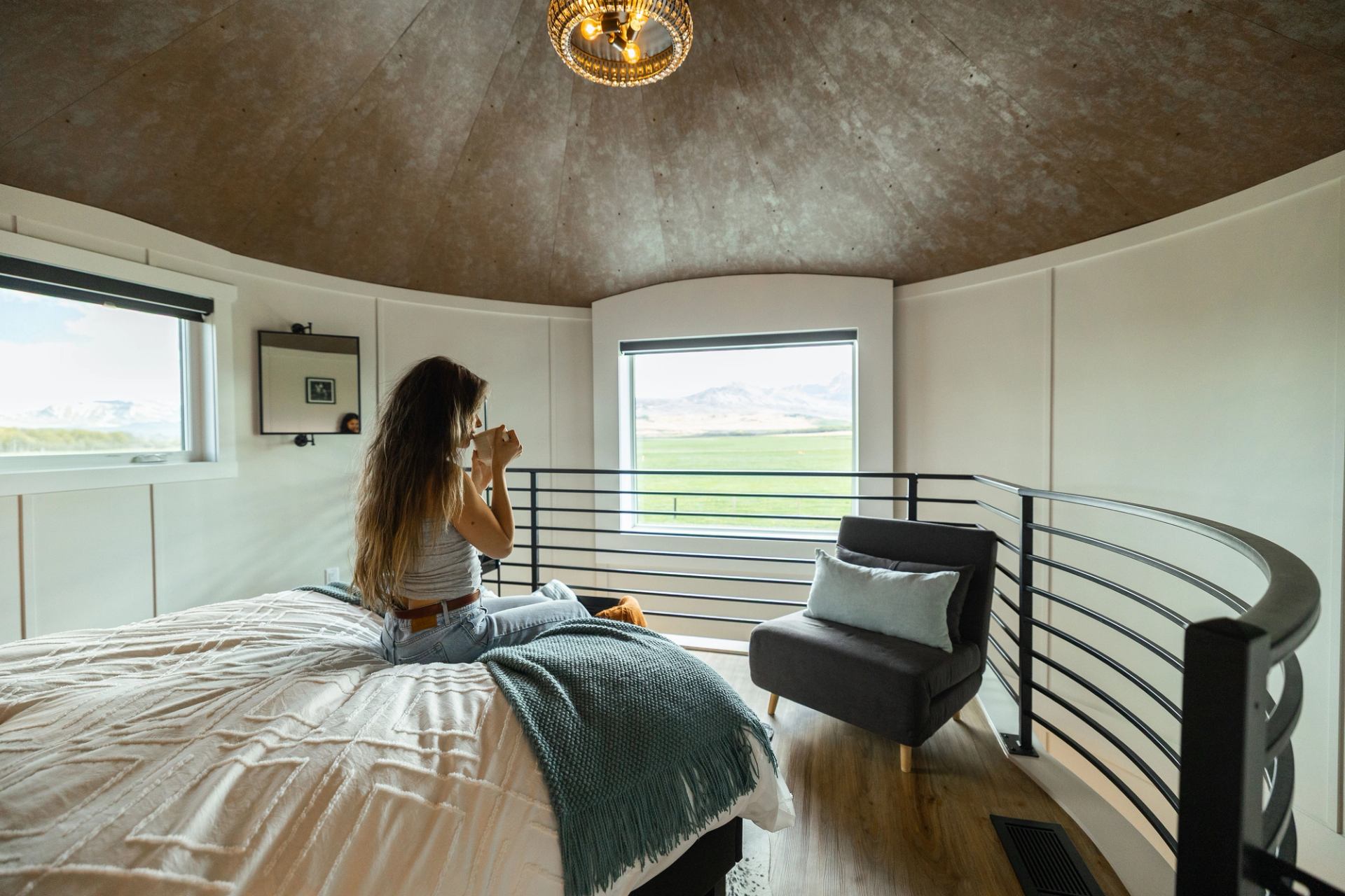 A woman sits on a cozy bed in a loft room with curved sides, inside a converted grain bin hotel room at Twin Butte Silos.