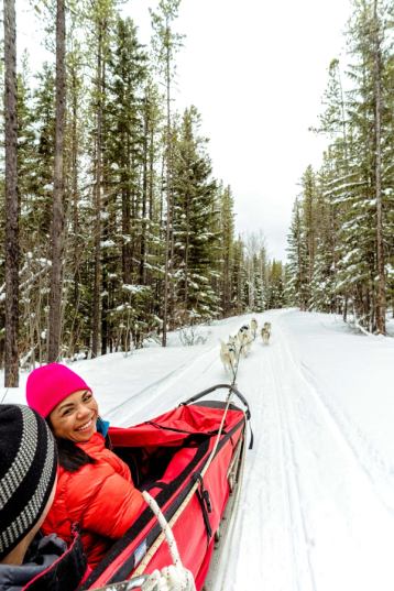 A women participant turns and smiles at the camera while on a dog sledding tour through a snow covered forest.