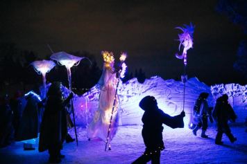 A group of performers walk down an icy path while holding light sticks at night.