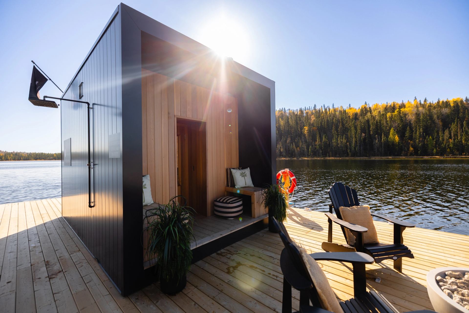 A floating sauna on a forested lake features a deck and chairs.
