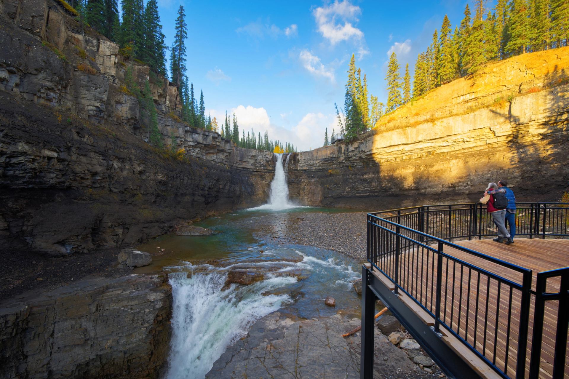 Two people stand on a platform viewing a two-tiered waterfall cascading into a rocky canyon.