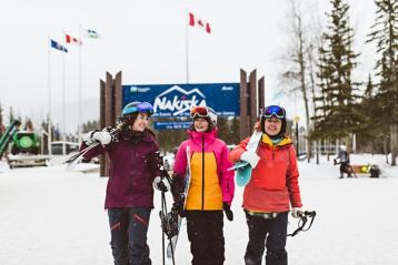 One female snowboarder and two female skiers smiling and walking in a line away from a ski resort sign.