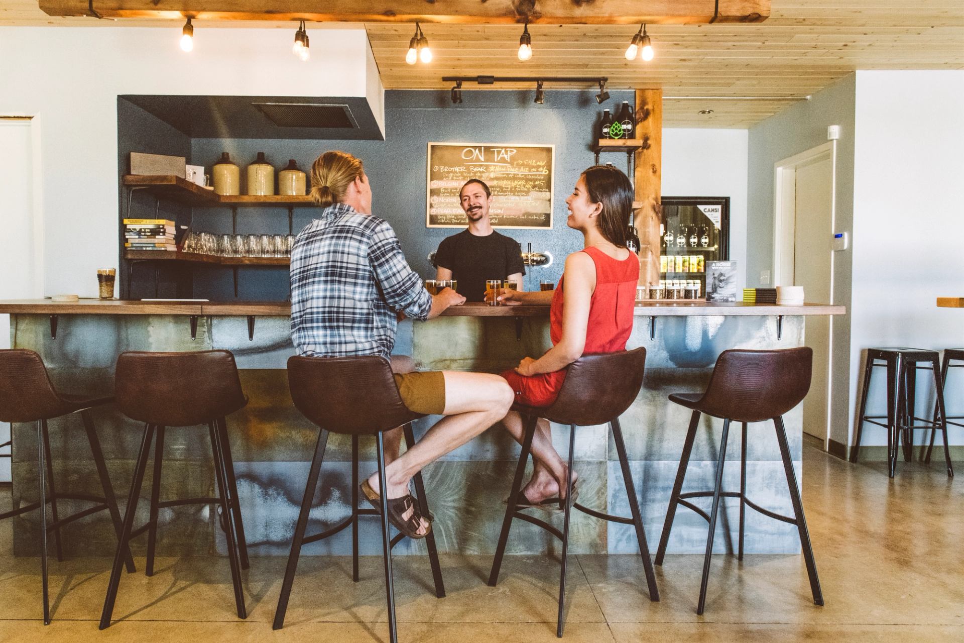 Two women sit at a brewery tasting bar with samples of beer while a bartender smiles at them.