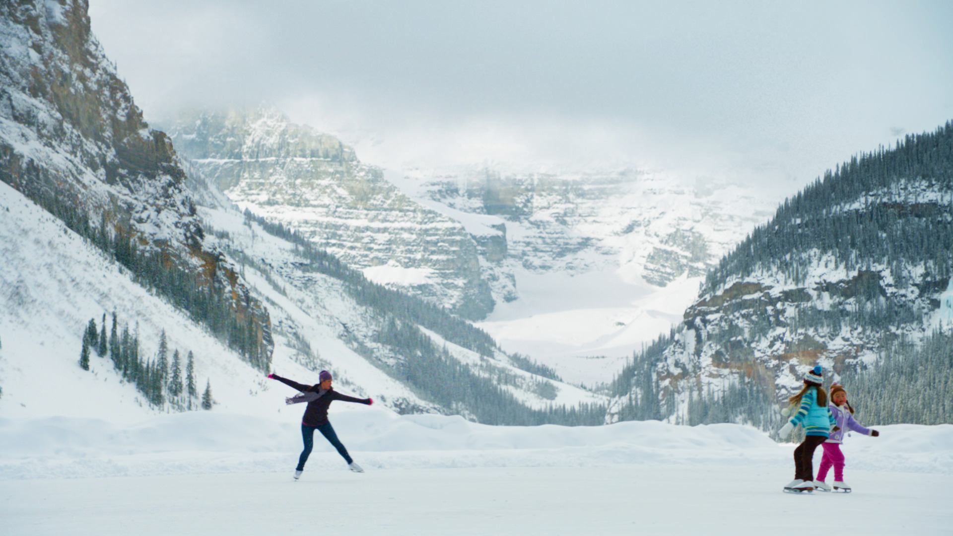 Family ice skating on a frozen lake with views of the mountains in the background.