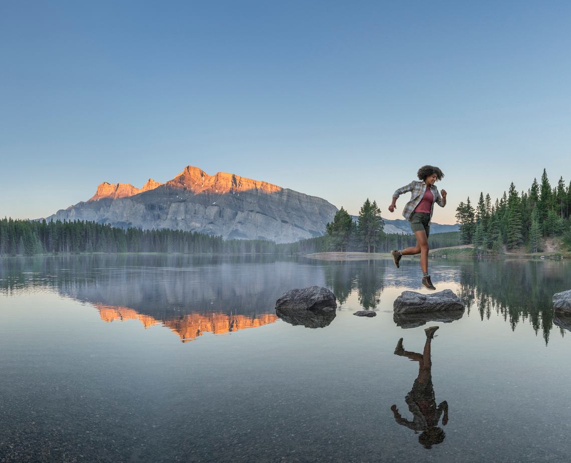 Woman jumping from rock to rock at on a lake with a mountain in the background.