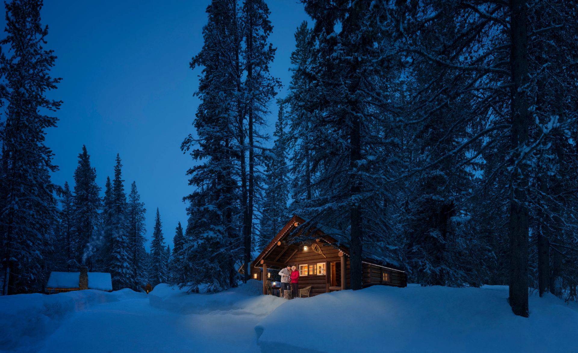 Couple drinking champagne at night outside a log cabin in the snowy trees at Storm Mountain Lodge