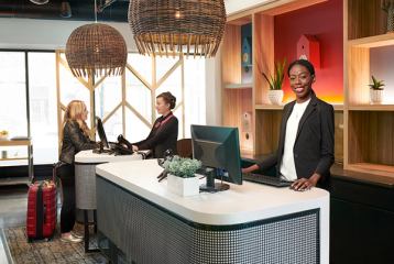 Receptionist smiling behind a modern reception desk while her colleagues checks in a guest with luggage beside her.