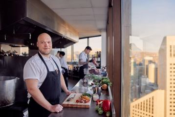 Chef in an apron posing by a chopping board by the window of a tall building with another chef in the background preparing food.