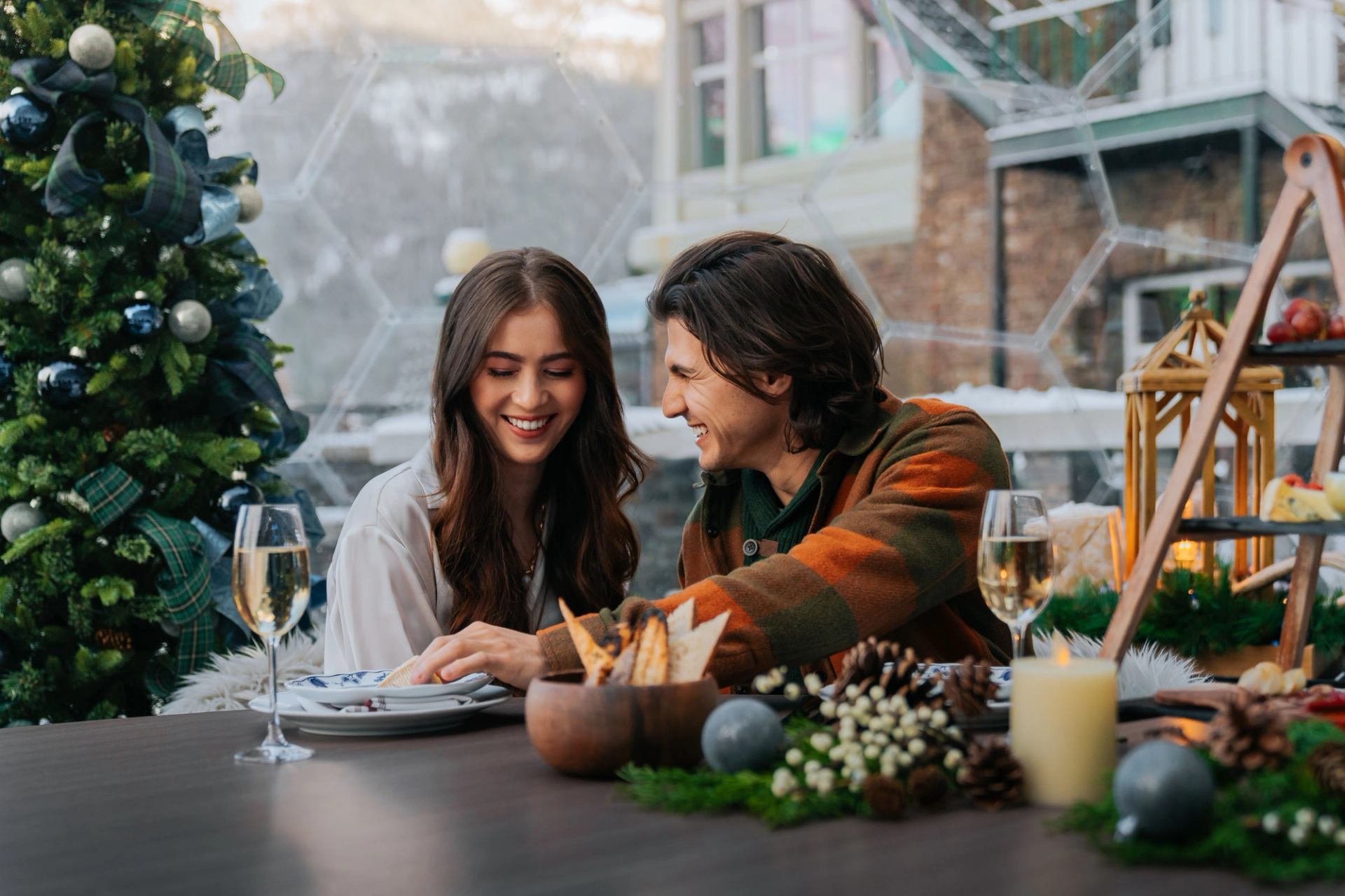 A couple sits inside a geodome at the Fairmont Banff Springs alongside a Christmas tree, food and glasses of champagne.