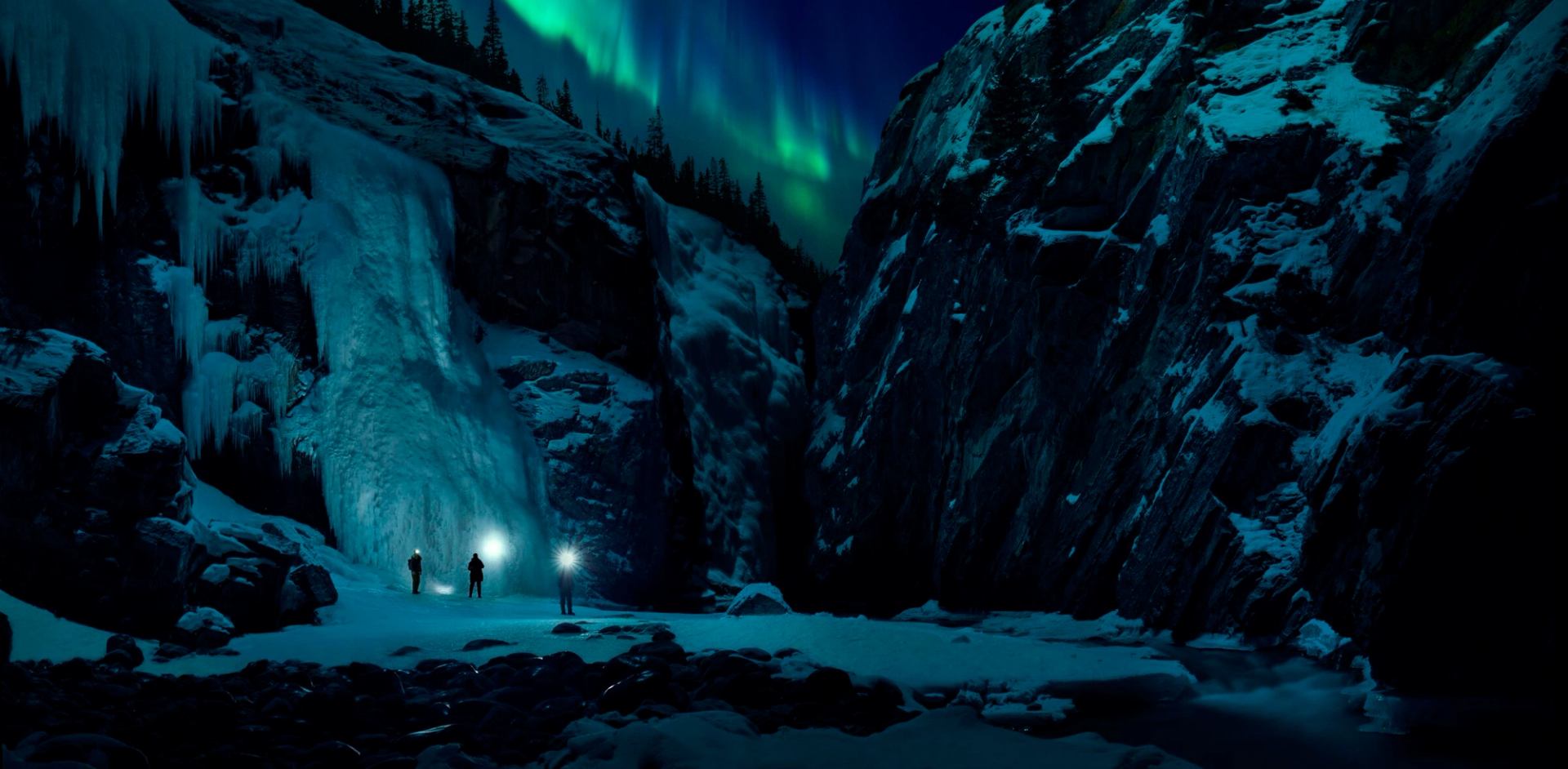 Group of three friends exploring a frozen canyon at nights with the Northern Lights overhead.
