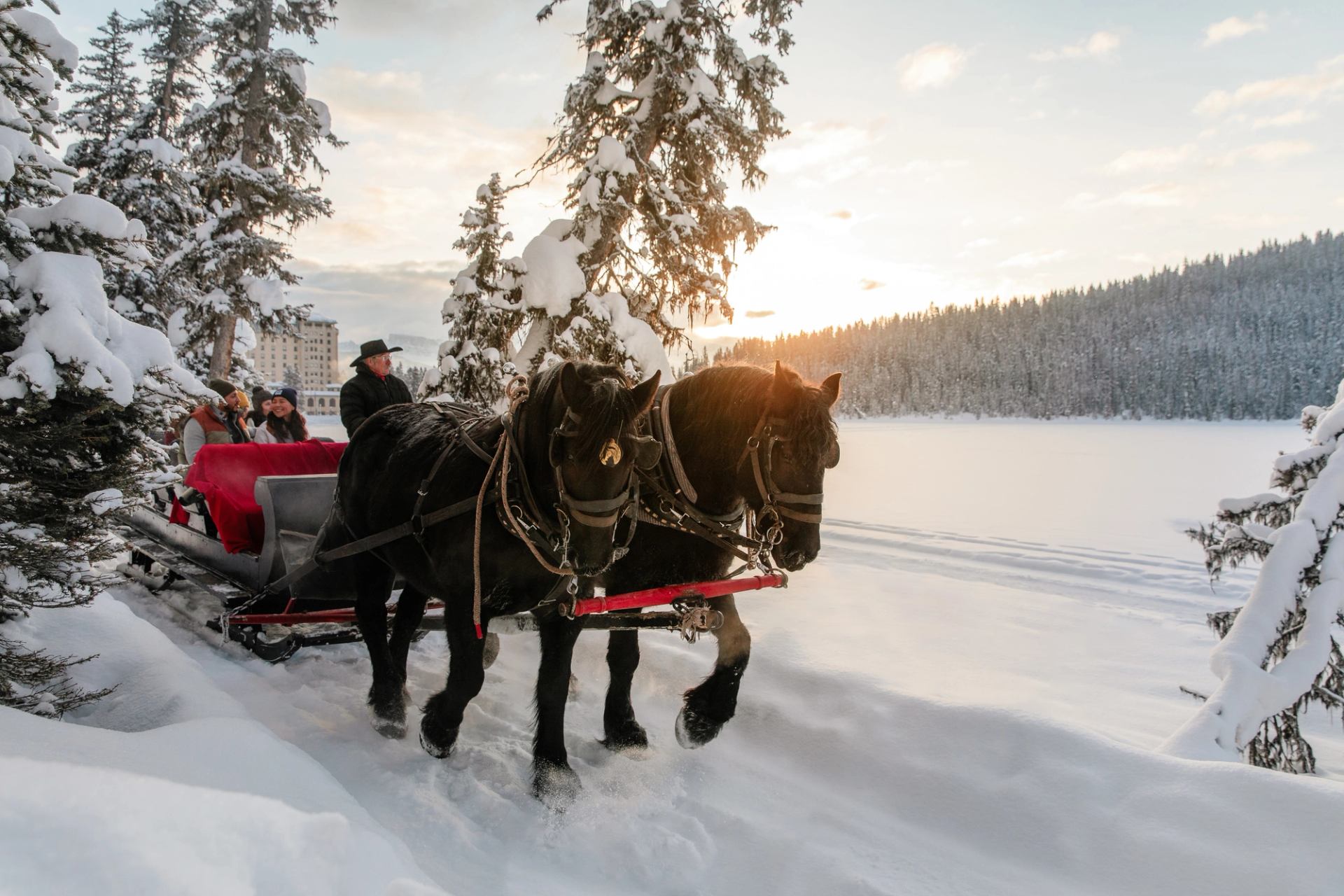 The sun rises behind a sleigh ride along the snowy path beside frozen Lake Louise on an idyllic winter day in Banff National Park.